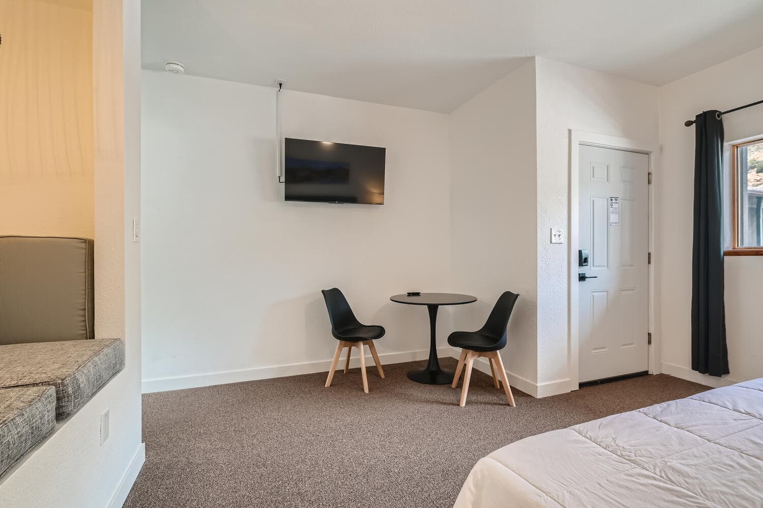 A small room featuring a round table with two chairs, a wall-mounted TV, and a door leading outside.