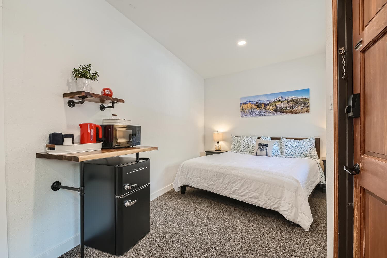 A cozy bedroom featuring a queen bed, kitchenette area, and a scenic wall art.