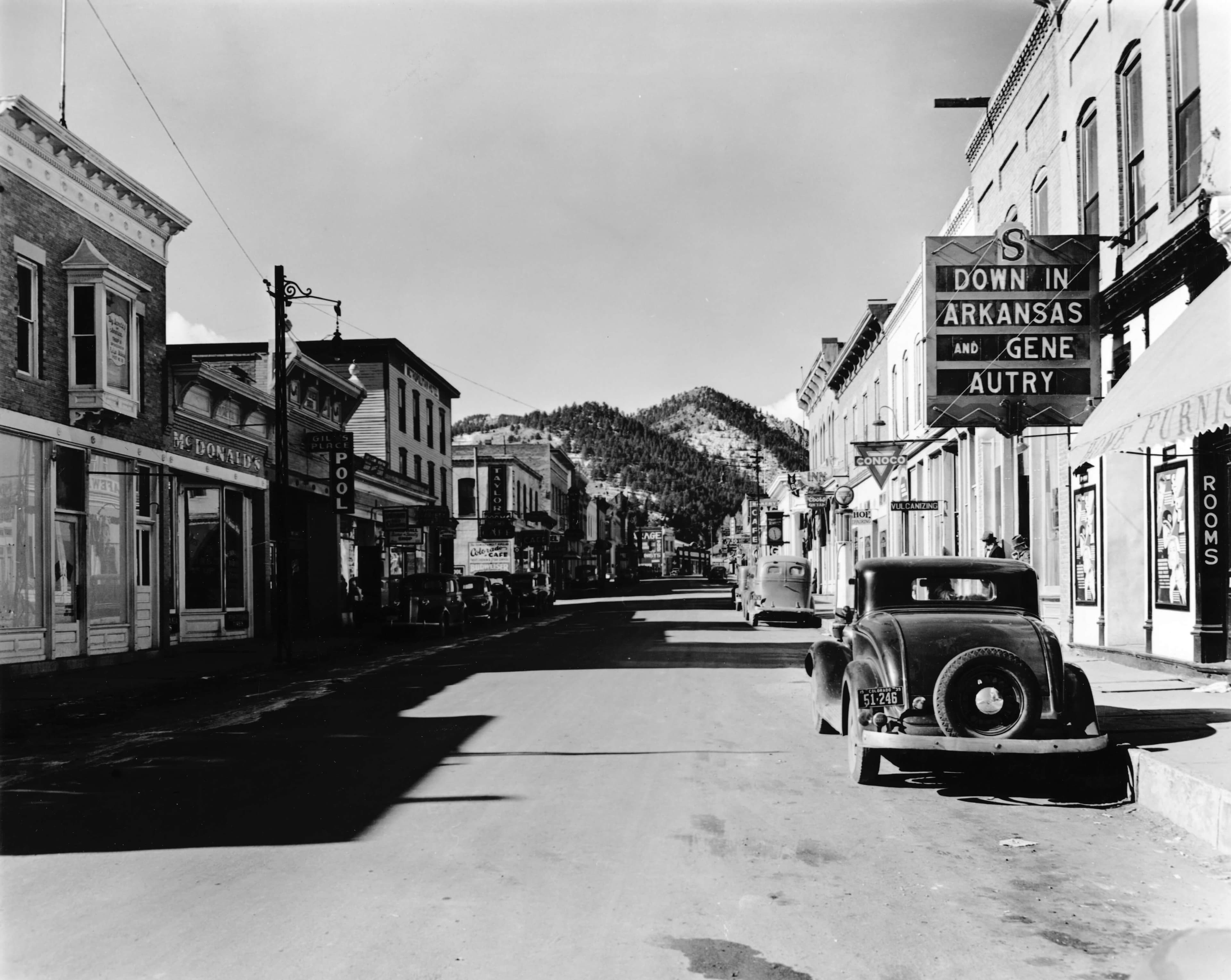 Black-and-white image of a quiet street lined with vintage storefronts and an old car parked in front.