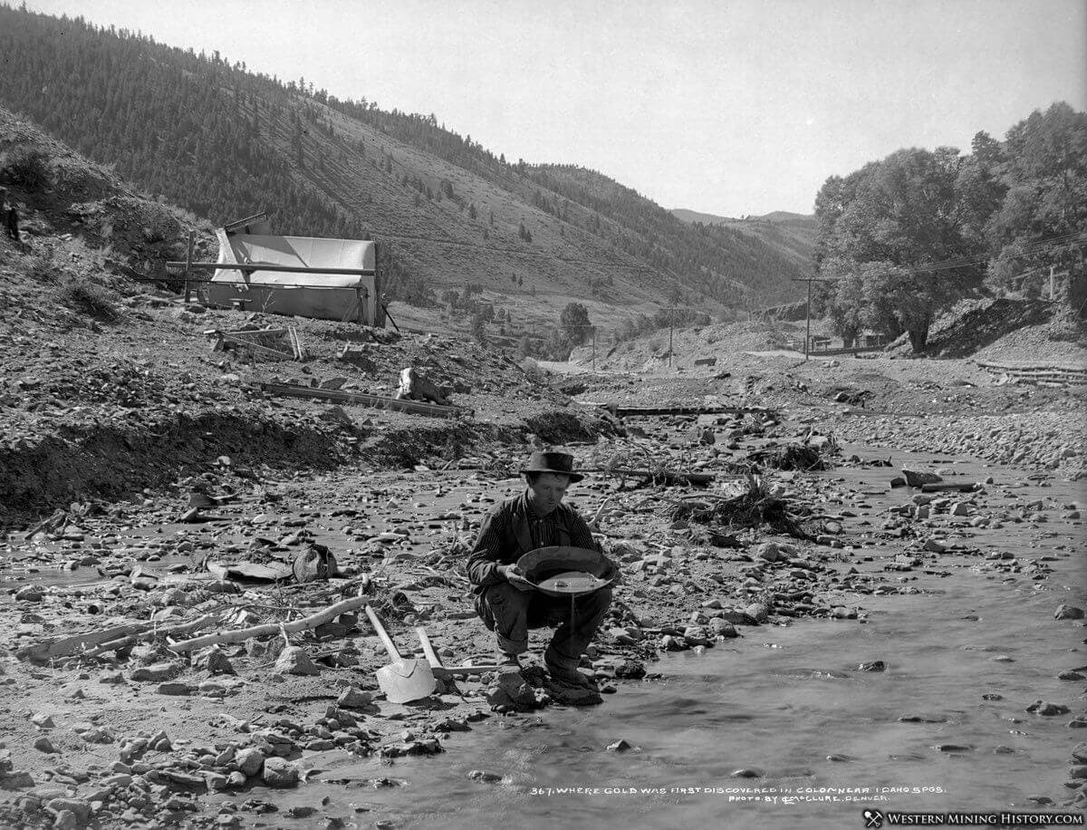 A miner pans for gold in a river surrounded by mountains and mining equipment.
