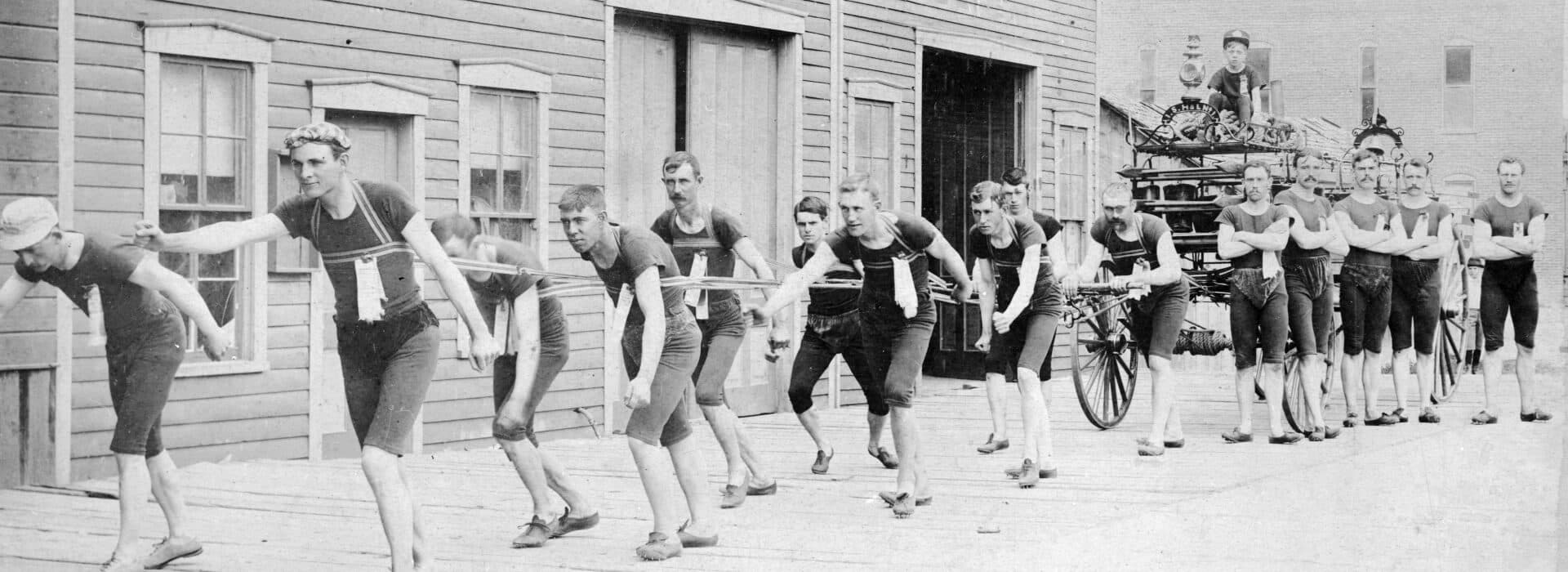 A group of men in matching outfits pulls a large cart along a wooden walkway.