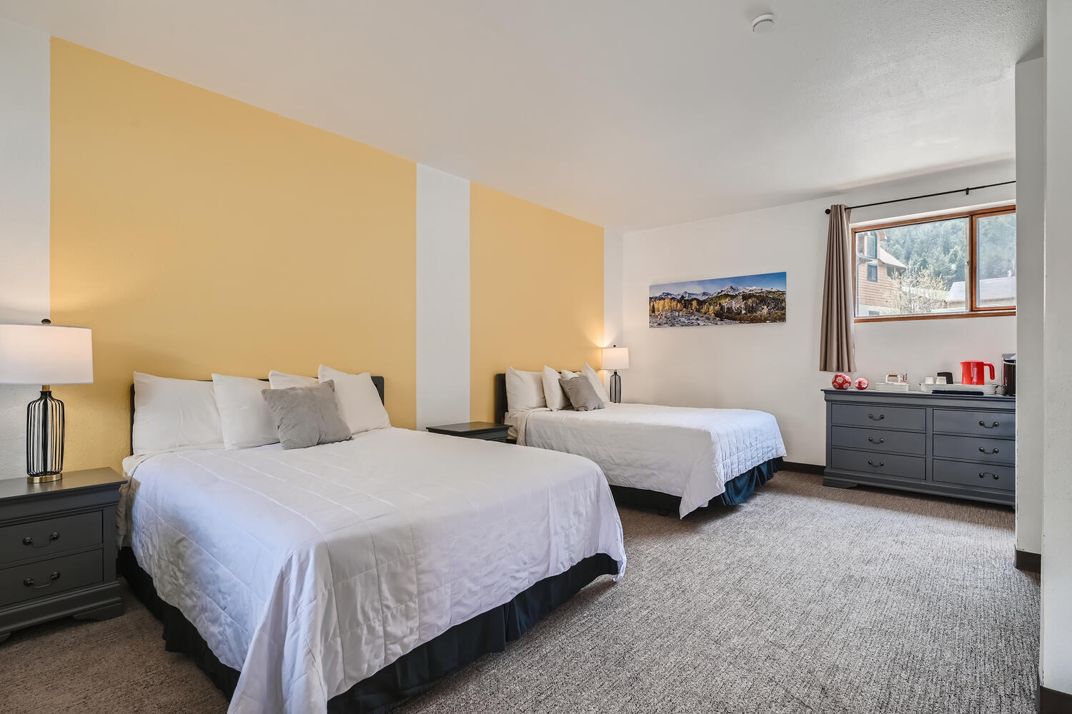 A brightly lit hotel room featuring two double beds, nightstands, and a window with a view.