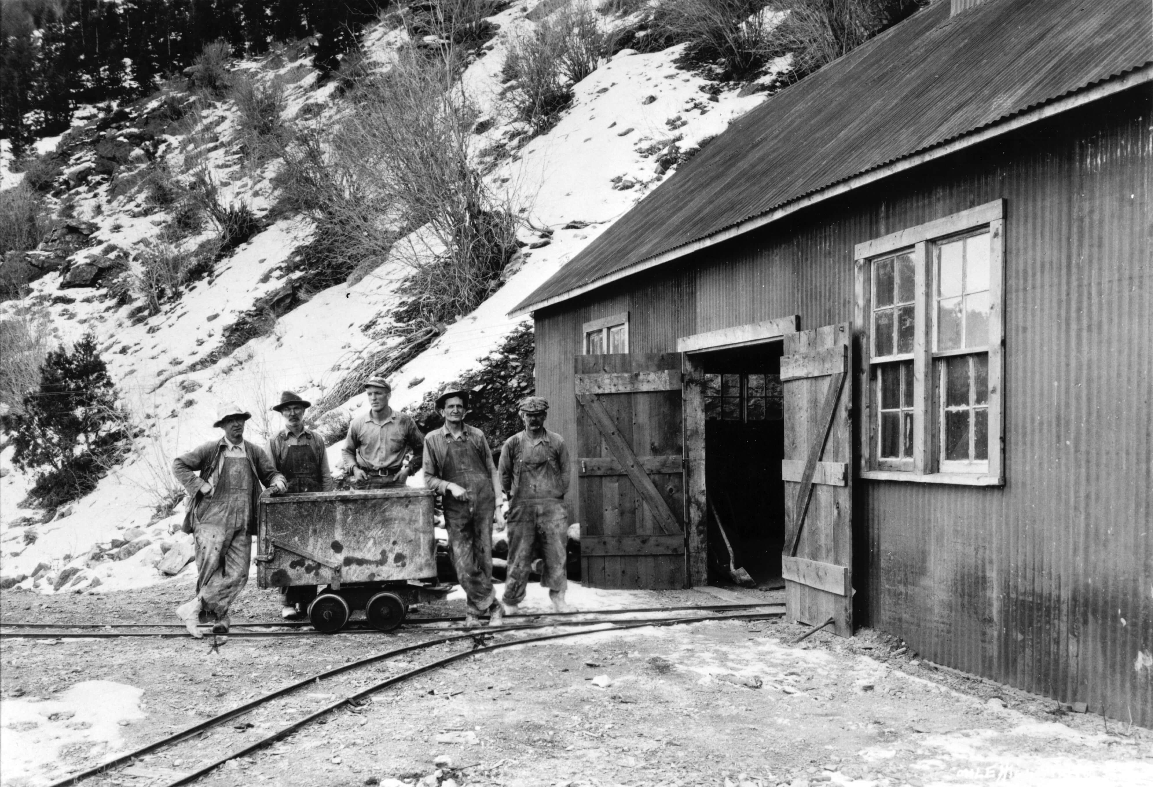 Five miners standing outside a wooden building, next to a small mine cart on tracks.