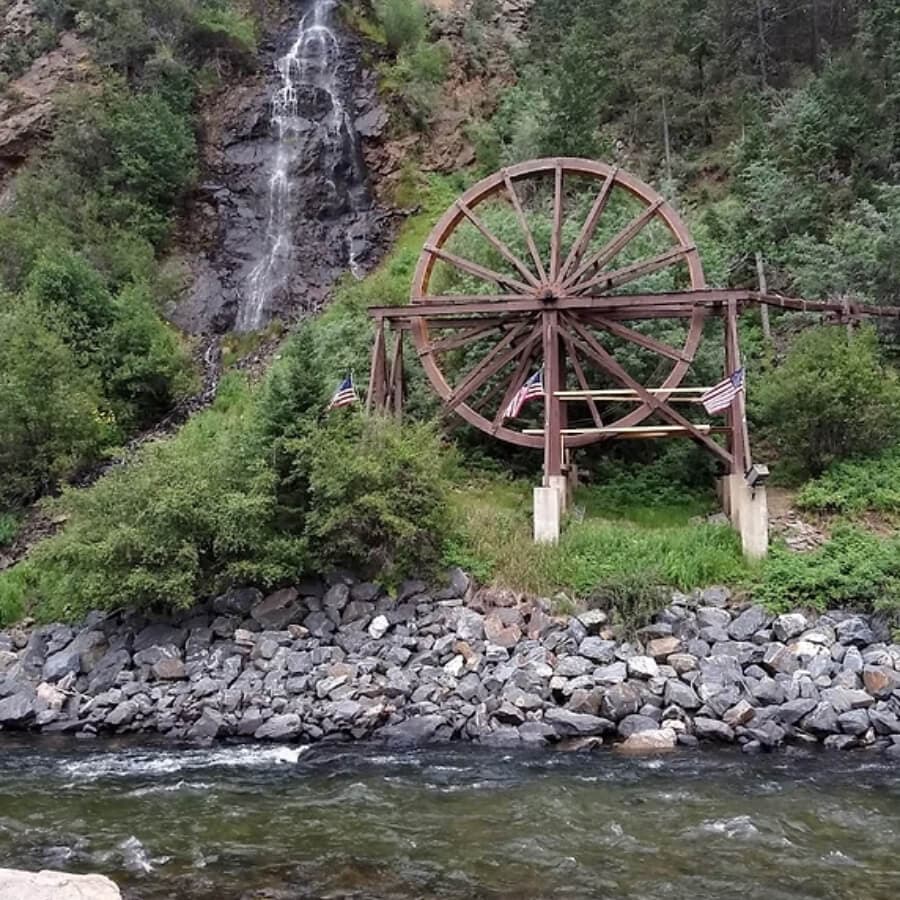 A wooden water wheel stands near a rocky riverbank with a waterfall in the background.