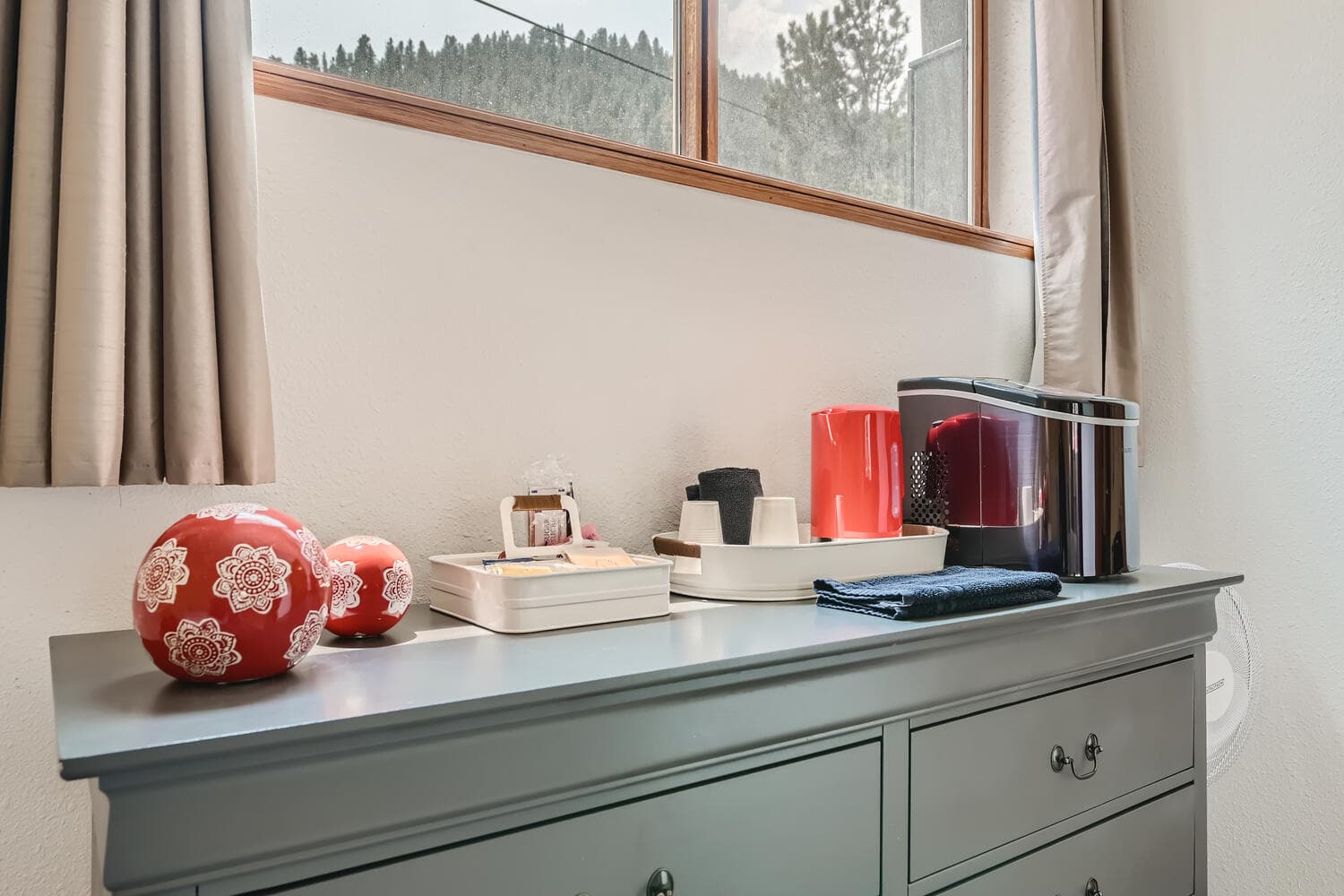 A grey dresser top adorned with decorative red vases, a coffee maker, and other kitchen items, beside a window with mountain views.