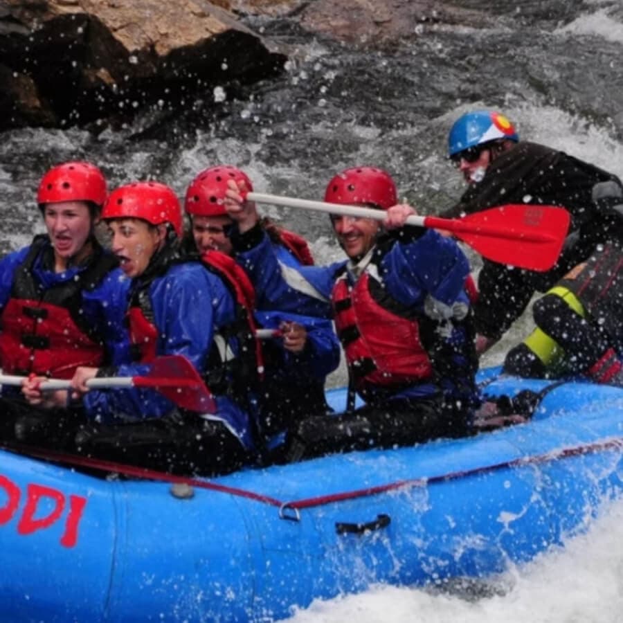 A group of six people in blue rafts and red helmets are whitewater rafting, splashing through turbulent water.