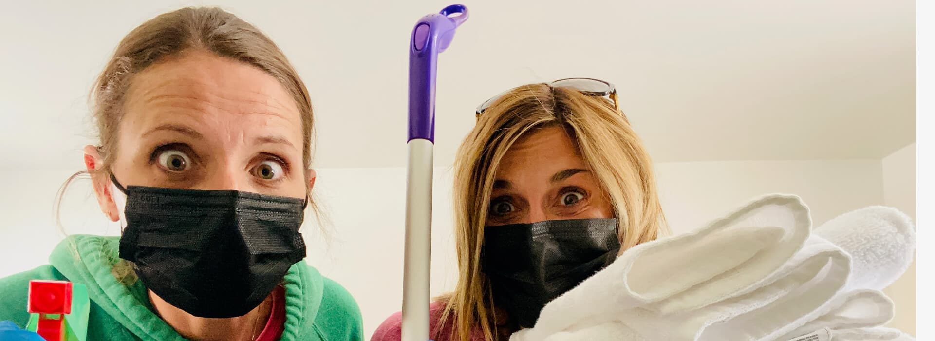 Two women wearing black masks express surprise while holding cleaning supplies and gloves.