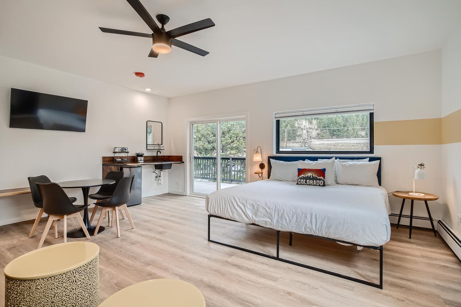 A modern bedroom with a bed, dining table, kitchenette, and large windows overlooking greenery.