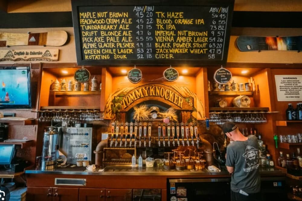 A person stands at a wooden bar with a large selection of beer taps and a menu board displaying various brews.