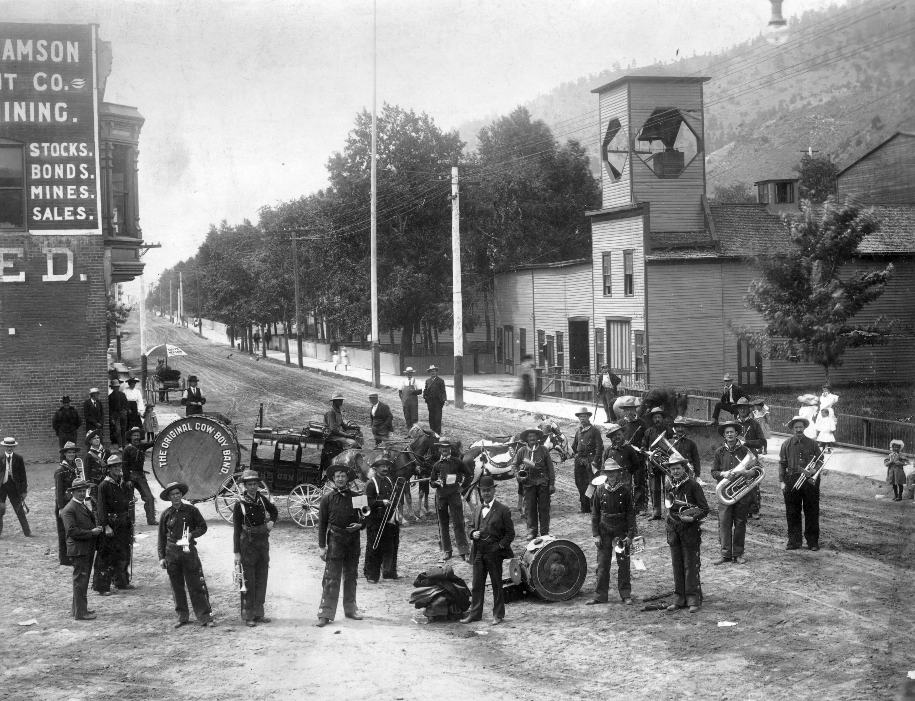 A vintage black-and-white photograph of a brass band performing on a city street, surrounded by spectators and buildings.