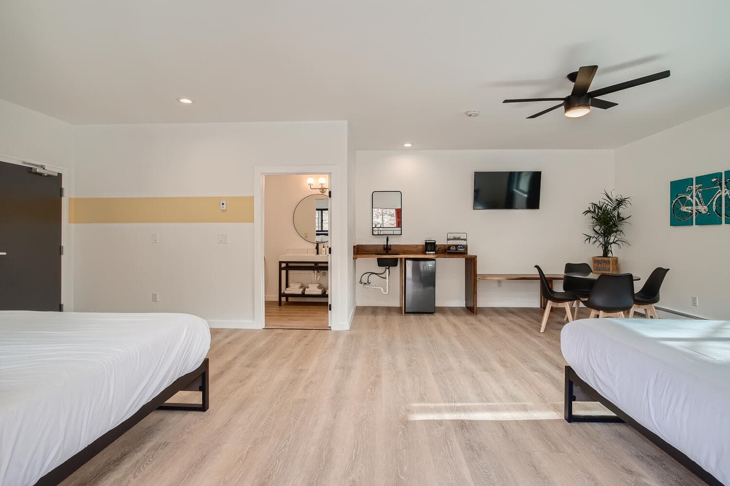 A spacious hotel room with two beds, a work desk, a TV, and a bathroom doorway.