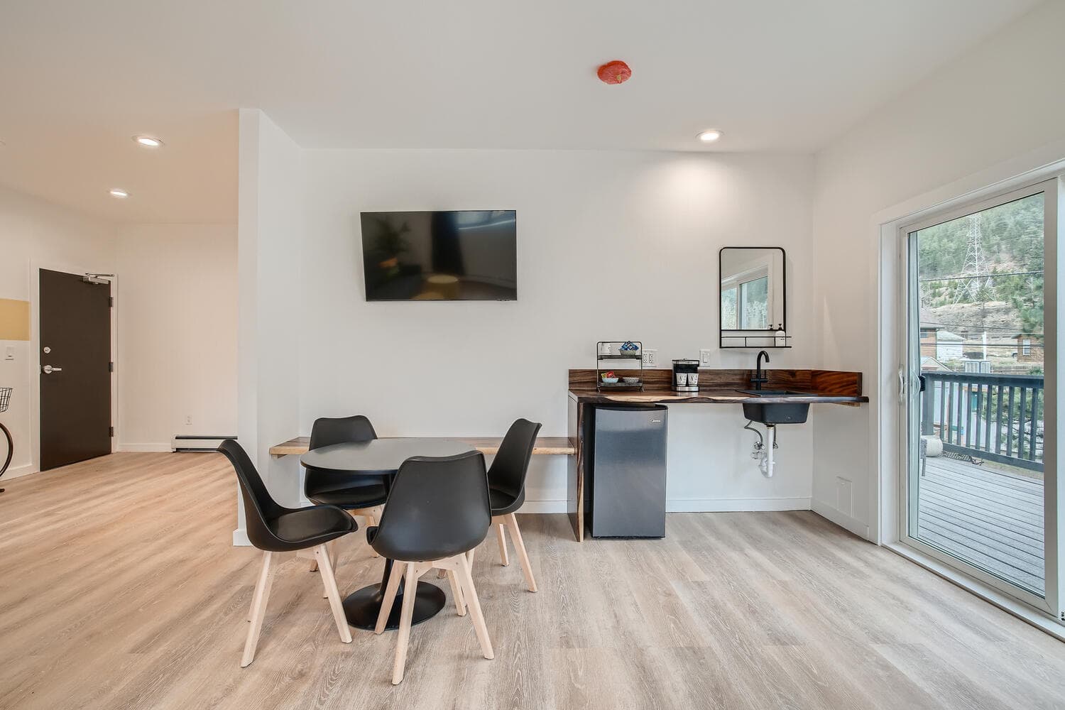 A modern kitchenette area featuring a table with black chairs, a coffee machine, and a wall-mounted TV.