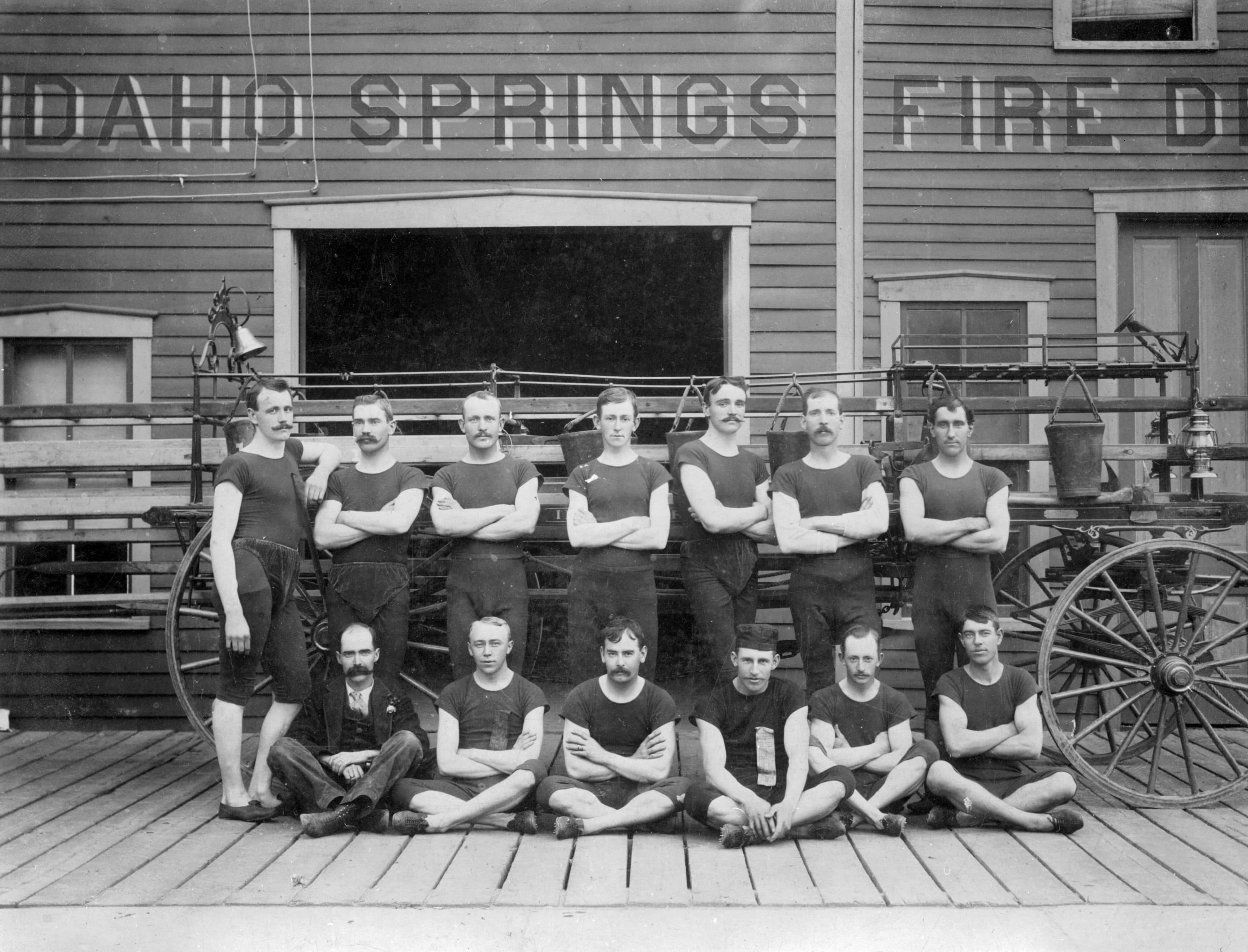 A historical black and white photograph of a group of eleven male firefighters posing in front of a fire truck outside the Idaho Springs Fire Department.